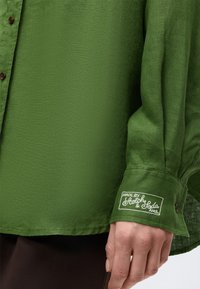 Πράσινο πουκάμισο με μακριά μανίκια και καφέ κουμπιά, ετικέτα στο μανίκι που γράφει «Made by Scotch & Soda AMS», φορεμένο από άτομο με χαλαρό χέρι.