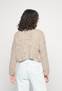 Suéter de ganchillo en beige, con patrones de encaje detallados, mangas largas y un dobladillo corto. Combinado con jeans de mezclilla azul claro.