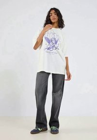 Hvid oversized T-shirt med lilla engel grafik og tekst, parret med mørkegrå vidde jeans og blå sneakers med grønne accenter.