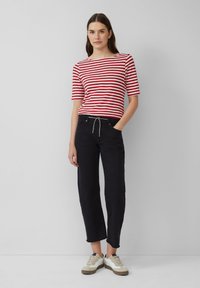 Camicia a righe rossa e bianca a maniche corte, pantaloni neri a gamba dritta con cordoncino, e sneakers beige con dettagli scuri.