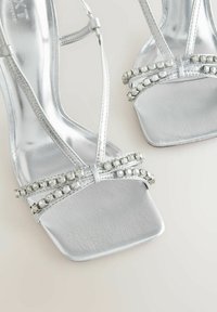 Stříbrné sandály na vysokém podpatku z lesklého materiálu, s plochými čtvercovými špičkami a dvojitými pásky zdobenými průhlednými rhinestones. Hladká textura a elegantní design.