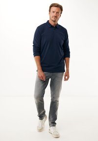 Polo shirt a maniche lunghe blu navy con un colletto a tre bottoni, abbinato a jeans grigi chiari e sneakers bianche su uno sfondo neutro.