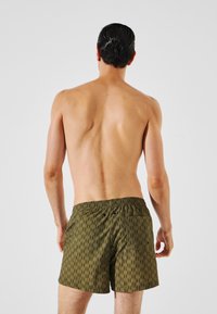 Costumi da bagno da uomo in verde scuro con un motivo geometrico. Presentano un elastico in vita e una tasca posteriore con due chiusure a bottone.