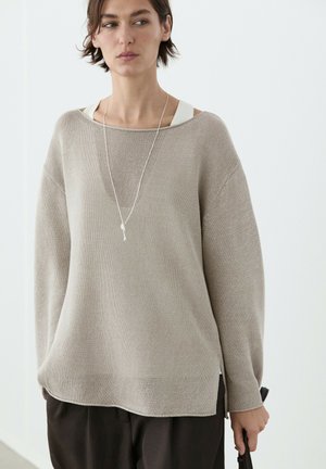 Frau trägt einen locker sitzenden beigen Strickpullover über einem weißen Top, schwarze Hose und lange silberne Halskette, hält einen schwarzen Gegenstand in der rechten Hand.