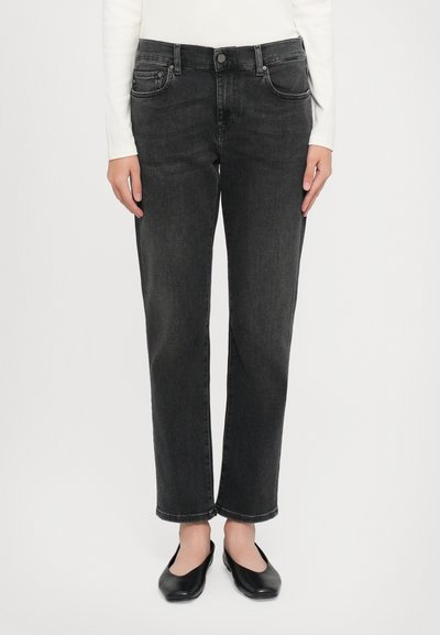 Mujer vestida con jeans rectos de talle alto negros, blusa blanca de manga larga y zapatos planos negros, de pie contra un fondo blanco.