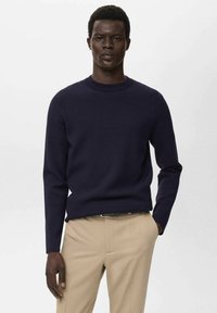 Navyblauer Strickpullover mit Rundhalsausschnitt, langen Ärmeln und gerippten Bündchen, kombiniert mit beigen Hosen. Strukturierte Stoffoberfläche.