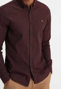 Hombre que lleva una camisa de botones de color rojo oscuro con un pequeño logo bordado amarillo en el pecho y pantalones beige, con las manos en los bolsillos.