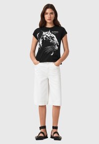 Camiseta negra de algodón con un gráfico de tigre, combinada con pantalones cortos de denim blancos y sandalias negras con correas en el tobillo y acentos de hardware dorado.