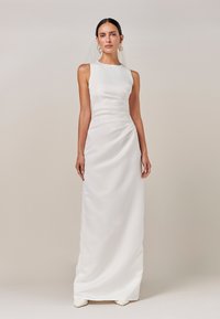 Lång, vit satinklänning med figurnära silhuett, ärmlös design, subtil drapering vid midjan och en slät textur, kombinerad med spetsiga skor.