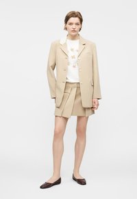 Γυναίκα που φοράει μπεζ σακάκι και πλισέ φούστα, λευκό τοπ με λουλούδια και σκούρα καφέ φλατ παπούτσια, στέκεται μπροστά σε απλό φωτεινό φόντο.