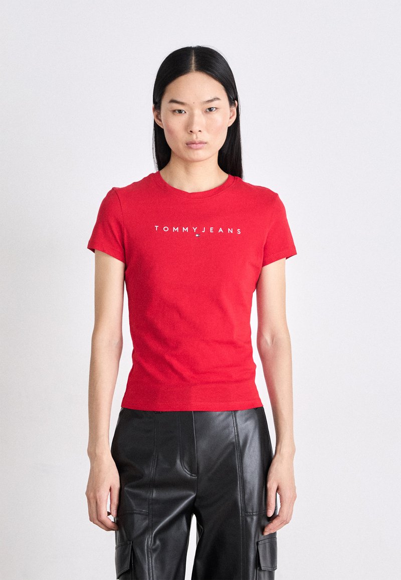 Červené bavlněné tričko s krátkými rukávy, kulatým výstřihem a bílým logem "TOMMY JEANS" umístěným na hrudi. Model má na sobě černé kožené kalhoty.