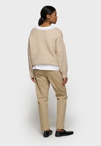 Beige gebreide trui met een relaxed fit, over een wit shirt gedragen; gecombineerd met beige broeken en zwarte schoenen. Soepele textuur, eenvoudig ontwerp.