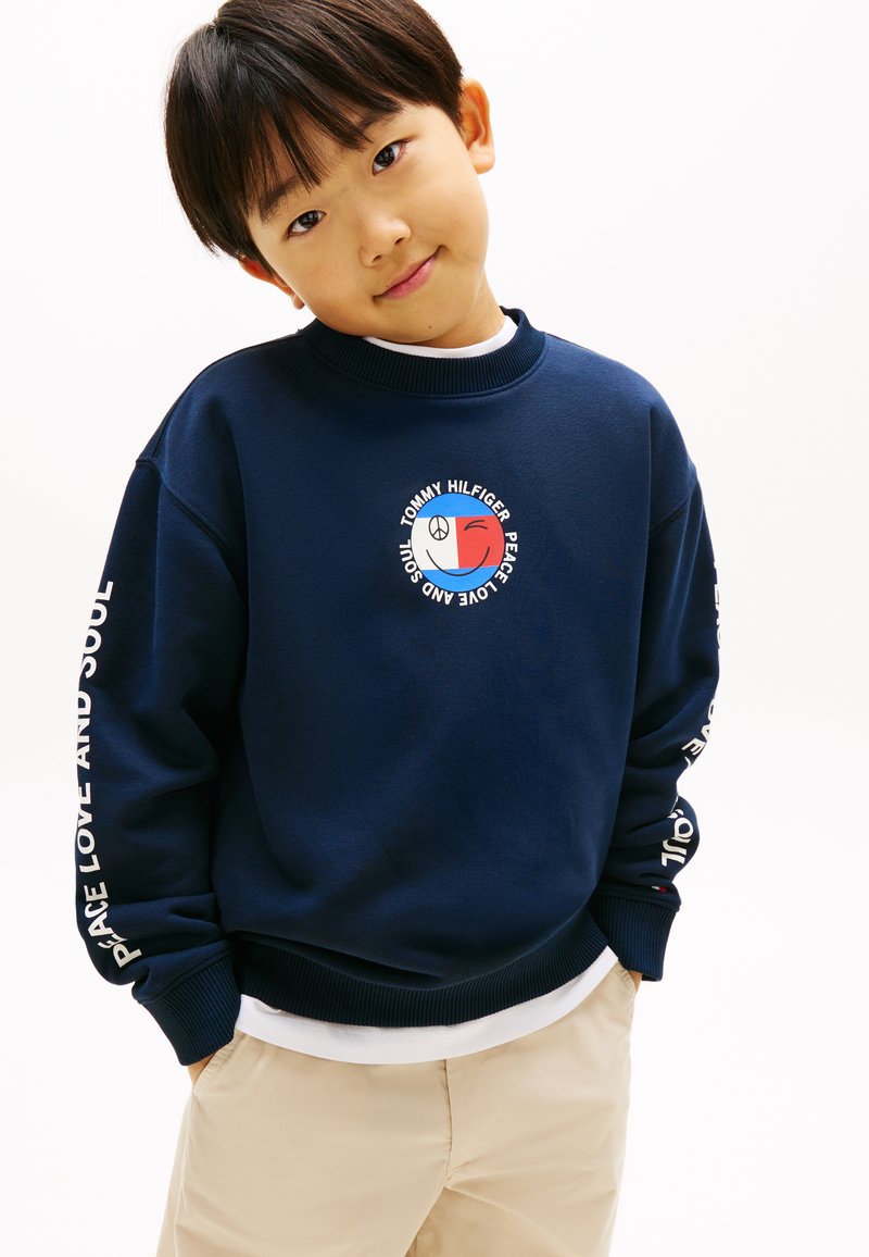 Νεαρός αγόρι που φοράει μπλε σκούρο φούτερ με κείμενο και γραφικά «Tommy Hilfiger Peace Love and Soul», μπεζ παντελόνι, χέρια στις τσέπες, ελαφριά κλίση του κεφαλιού.