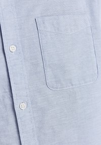 Ljusblå skjorta med knappar i ett tyg med subtil textur, med en bröstficka och vita knappar längs knappraden.