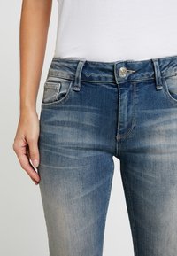 Jeans de mezclilla azul desteñido con una cintura de altura media, bolsillos frontales y cierre de botón metálico. Material elástico con una textura suave.