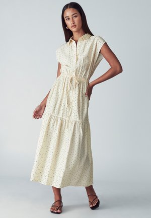 Długa, kremowa sukienka z krótkimi rękawami, ze sznurkiem w talii i czarnymi kropkami. Posiada zapięcie na guziki z przodu oraz falbaniasty dół.