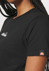 Personne portant un t-shirt noir à manches courtes avec le logo blanc "ellesse" sur la poitrine et un petit logo coloré sur la manche, cheveux tressés visibles.