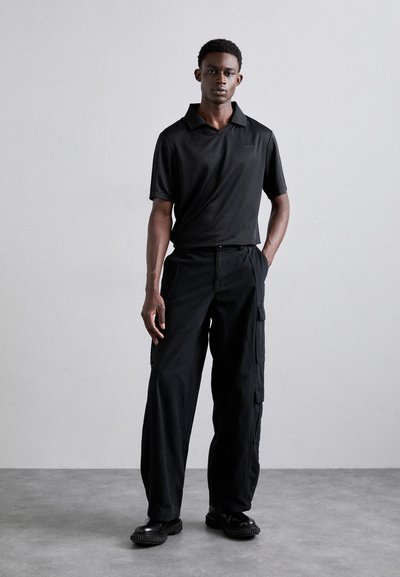 Svart polo skjorte med krage og korte ermer, kombinert med svarte cargo bukser med sidelommer og en avslappet passform. Svarte sko og ensfarget bakgrunn.