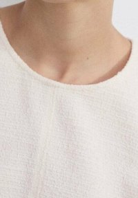 Haut texturé couleur crème avec un col rond, présentant un motif tissé subtil et des bords lisses, conçu sans fermetures visibles.