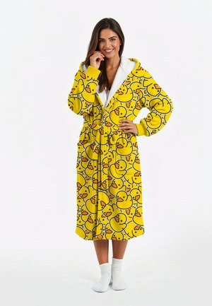 Naeratav naine kollase kapuutsiga hommikumantlis, mis on kummist pardimustriga, valge sokkides, seisab ühevärvilise valge tausta ees.