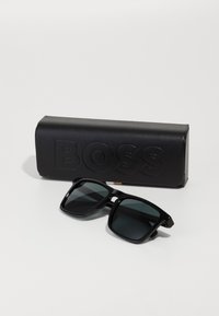 Óculos de sol pretos com lentes escuras e armações grossas, acompanhados por um estojo de couro preto em relevo com a marca "BOSS". Textura suave, forma retangular.