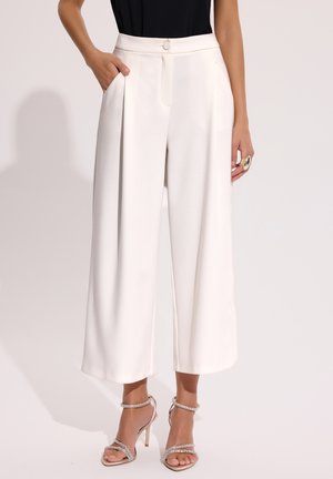 Kobieta ubrana w białe, plisowane spodnie z wysokim stanem i szerokimi nogawkami, w połączeniu z czarną bluzką i sandałami na wysokim obcasie z ozdobnym paskiem z cyrkonii.
