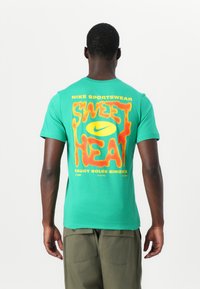 Grøn bomulds T-shirt med et stort orange og gult grafik på ryggen, der står "SWEET HEAT" med Nike logo og branding.