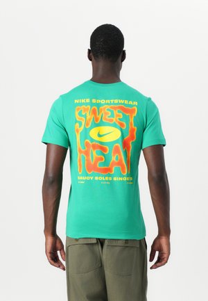 T-shirt verde de algodão com uma grande gráfica laranja e amarela nas costas que diz "SWEET HEAT" com o logótipo e a marca da Nike.