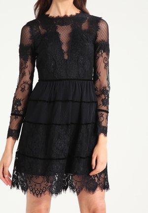 Frau trägt ein knielanges schwarzes Spitzenkleid mit transparenten langen Ärmeln und gewelltem Saum, verziert mit floraler Stickerei und tailliertem Schnitt.