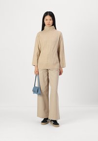 Bežs turtlenecks džemperis ar ribotu apakšmalu, kombinēts ar gaiši bežām platākām biksēm. Modelis tur mazu zilu somiņu; melni sporta apavi ar gumijas zoli.
