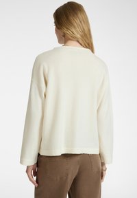 Frau mit langen blonden Haaren, die einen lockeren weißen Pullover und braune Hosen trägt, von hinten vor einem schlichten Hintergrund betrachtet.