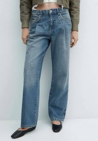 Mujer con jeans azules de cintura alta y sueltos, chaqueta corta de color oliva y zapatos planos negros de punta cuadrada, de pie contra un fondo claro y liso.