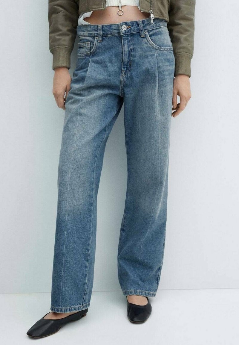 Mujer con jeans azules de cintura alta y sueltos, chaqueta corta de color oliva y zapatos planos negros de punta cuadrada, de pie contra un fondo claro y liso.