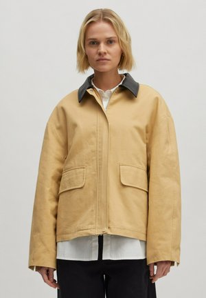 Beige oversized jas van stevige stof, met een zwarte kraag, twee voorzakken en een ritssluiting, getoond op een model.