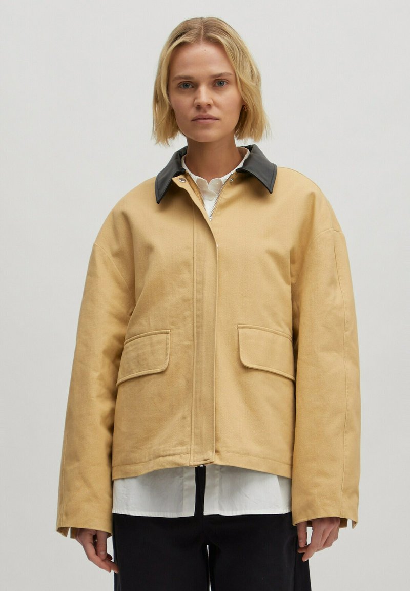 Beige Oversize-Jacke aus robustem Stoff, mit einem schwarzen Kragen, zwei Fronttaschen und einem Reißverschluss, auf einem Model präsentiert.
