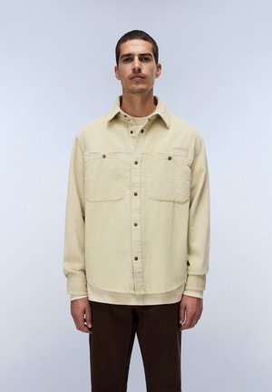 Man staat rechtop, draagt een lichtbeige corduroy overhemdjas en donkerbruine broek tegen een effen lichte achtergrond.