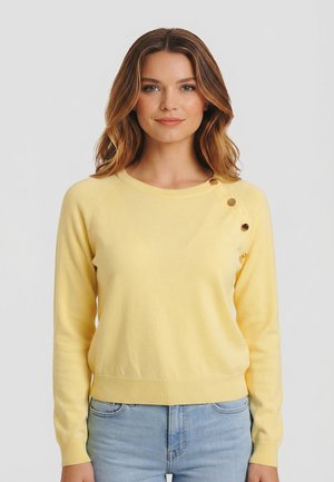 Mujer con un jersey de manga larga amarillo claro con tres botones dorados en el hombro izquierdo y vaqueros azul claro, de pie contra un fondo liso.