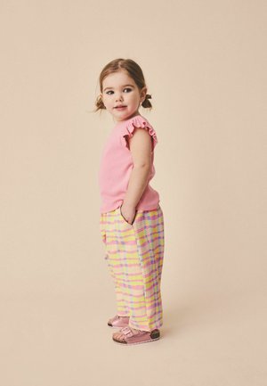 Kleines Mädchen mit braunen Haaren in Zöpfen trägt ein rosa Oberteil mit Rüschensäumen, gelb-rosa karierte Hosen und rosa Sandalen, steht mit den Händen in den Taschen.