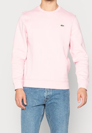 Mann trägt ein hellrosa Sweatshirt mit langen Ärmeln und kleinem grünem Krokodil-Logo sowie blaue Jeans, steht vor einem schlichten Hintergrund.