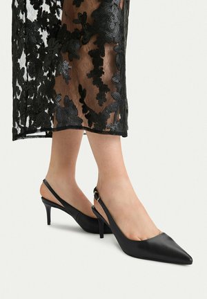 Nogi w przezroczystej czarnej spódnicy koronkowej oraz czarne czółenka na niskim obcasie typu slingback z paskami na kostce i bocznymi klamrami.