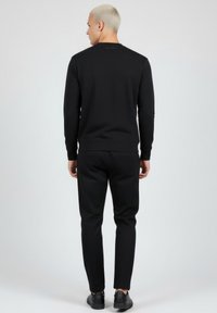 Sudadera negra con cuello redondo, mangas largas y puños acanalados. Combinada con pantalones negros a juego y zapatos negros. Detalle minimalista de logo en la parte trasera.