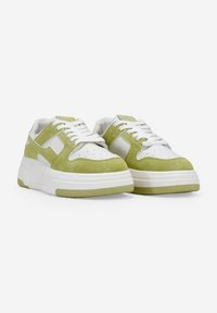 Zapatillas verdes y blancas con una suela gruesa, parte superior lisa y puntera perforada. Presentan cordones blancos y paneles laterales en ángulo.
