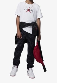 Vit t-shirt med svart "JORDAN"-tryck och röd logotyp, svart och röd jacka knuten i midjan, svarta byxor med zigzag-mönster, vita sneakers, röd väska.