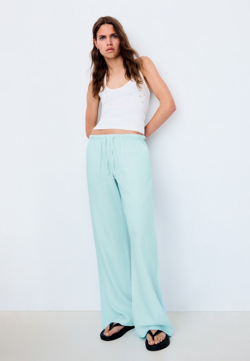 Femme portant un haut blanc sans manches, un pantalon large bleu clair et des tongs noires, debout devant un fond blanc uni.