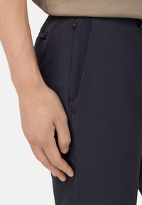 Tumed püksid sileda tekstuuriga, küljetaskute ja tõmbluku sulgemisega. Käsi toetub reie piirkonda, näidates kangaste üksikasju.