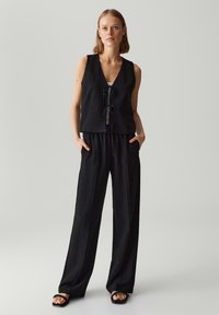 Zwarte mouwloze vest met een diepe V-hals en voorste knoopdetails, gedragen met losse zwarte broeken. Glad, lichtgewicht stof. Eenvoudig ontwerp.