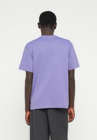 Man draagt een effen lavendelkleurig T-shirt met korte mouwen en donkere grijze broek, gezien van achteren tegen een witte achtergrond.