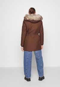 Žena v hnědé parce s kožíškem lemovanou kapucí, modrých džínách a černých botách stojí čelem k jednobarevnému bílému pozadí.