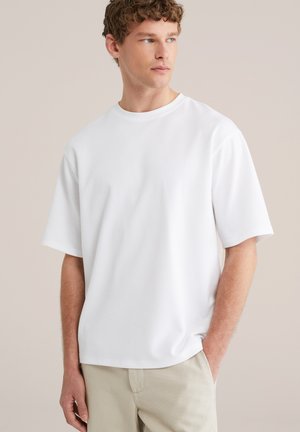 Fiatal férfi göndör hajjal, egyszerű fehér rövid ujjú pólót és bézs nadrágot visel, oldalra néz, egyik kezét a zsebében tartva.