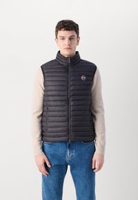 Gilet imbottito nero con colletto alto, chiusura con zip e tasche laterali. Presenta un patch con logo sul lato sinistro del petto, abbinato a un maglione leggero.